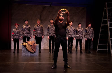 Antony all in black and wearing sunglasses holds up the laurel wreath crown as the other acts, wearing camoflaged shirts and black pants stand in a line in front of the marble backdrop with a ladder to the right.